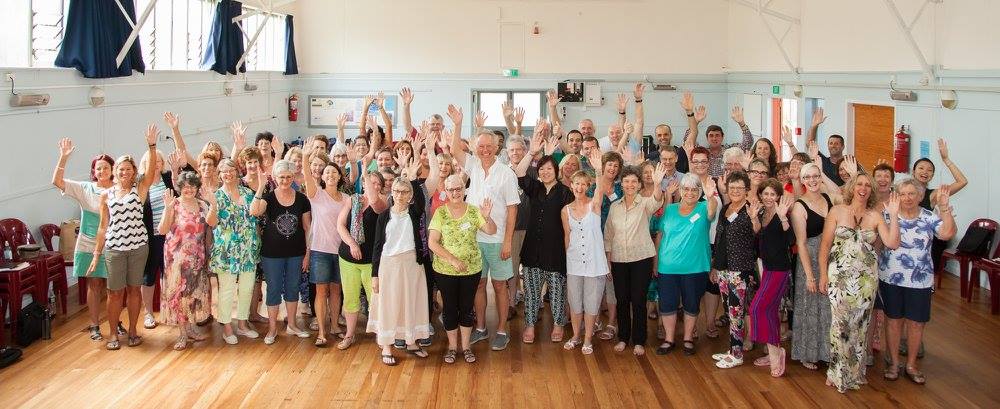 Picture of about 40 Auckland Choral singers, with its Music Director Uwe Grodd, in casual/relaxed clothes, in a venue on Waiheke Island, waving at you